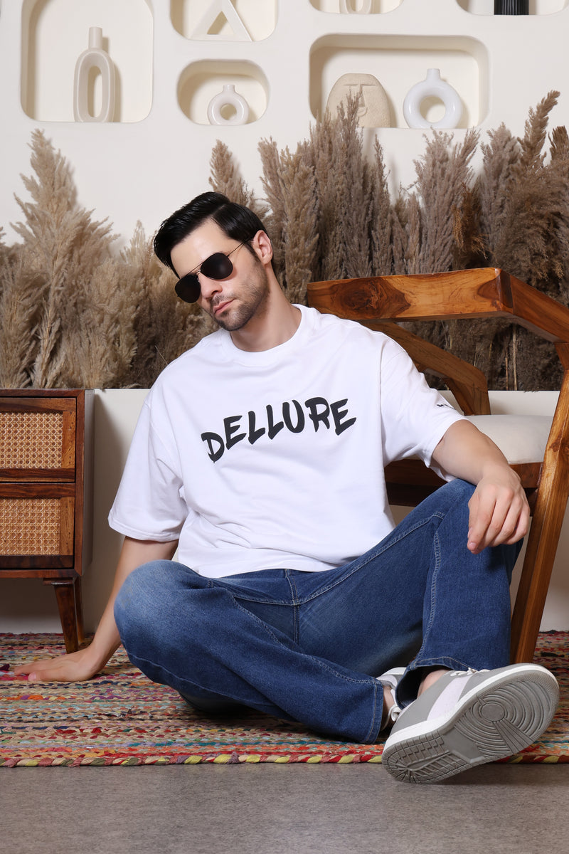 Man sitting on a wooden chair wearing a white t-shirt with 'DELLURE' text, surrounded by decorative plants.
